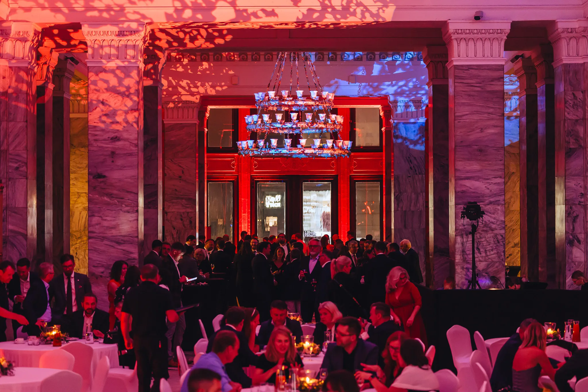 A hall with marble columns bathed in red light during a gala in Warsaw, guests at the tables and a chandelier above the entrance