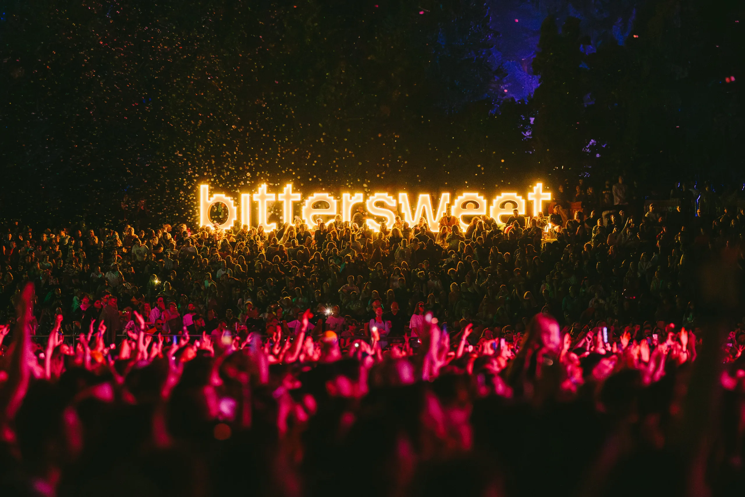 A crowd in front of the stage at a music festival at night, with raised hands and an illuminated sign above the audience