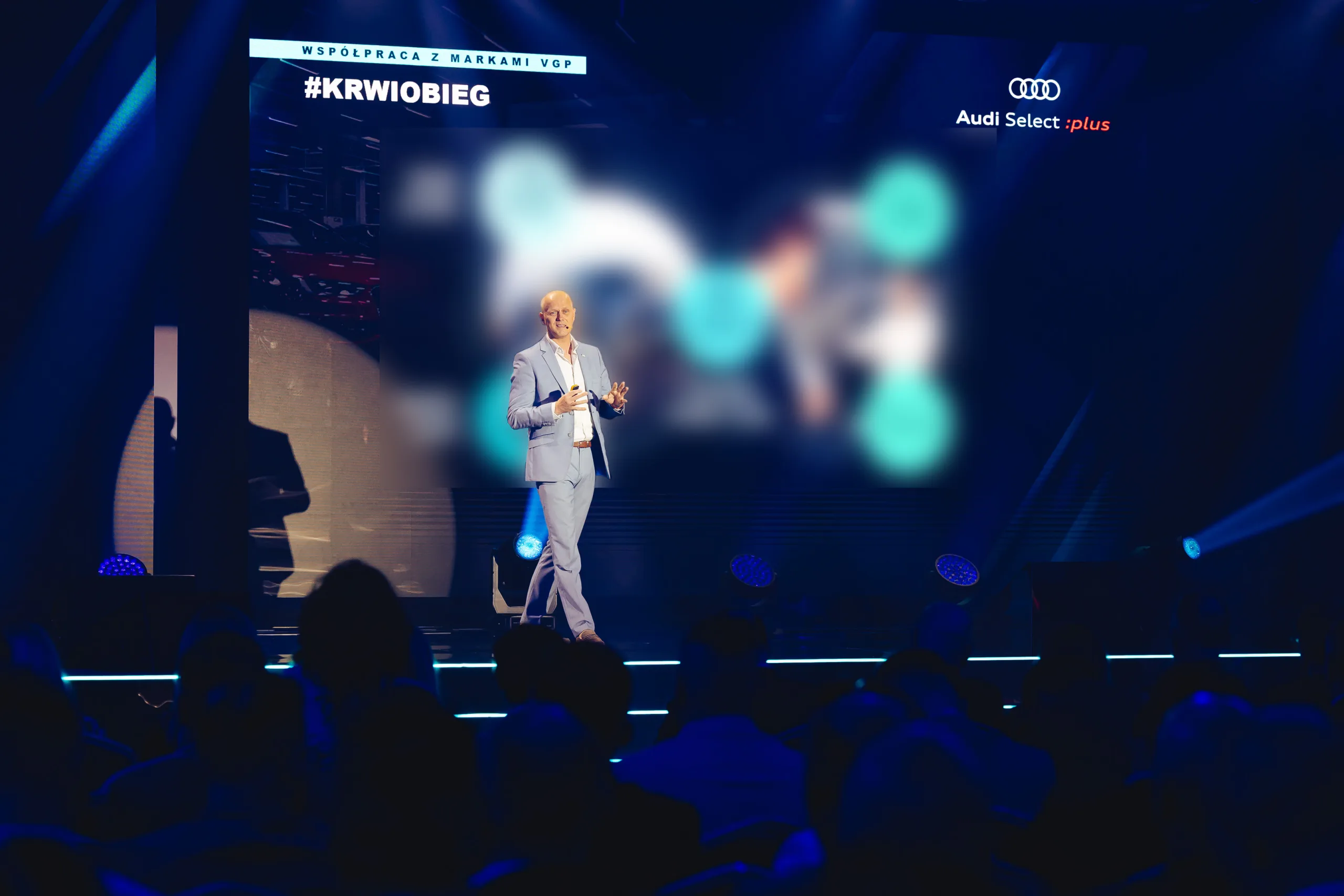 A speaker on the stage at a corporate conference in Warsaw, with the audience bathed in blue light in front of a screen displaying a presentation