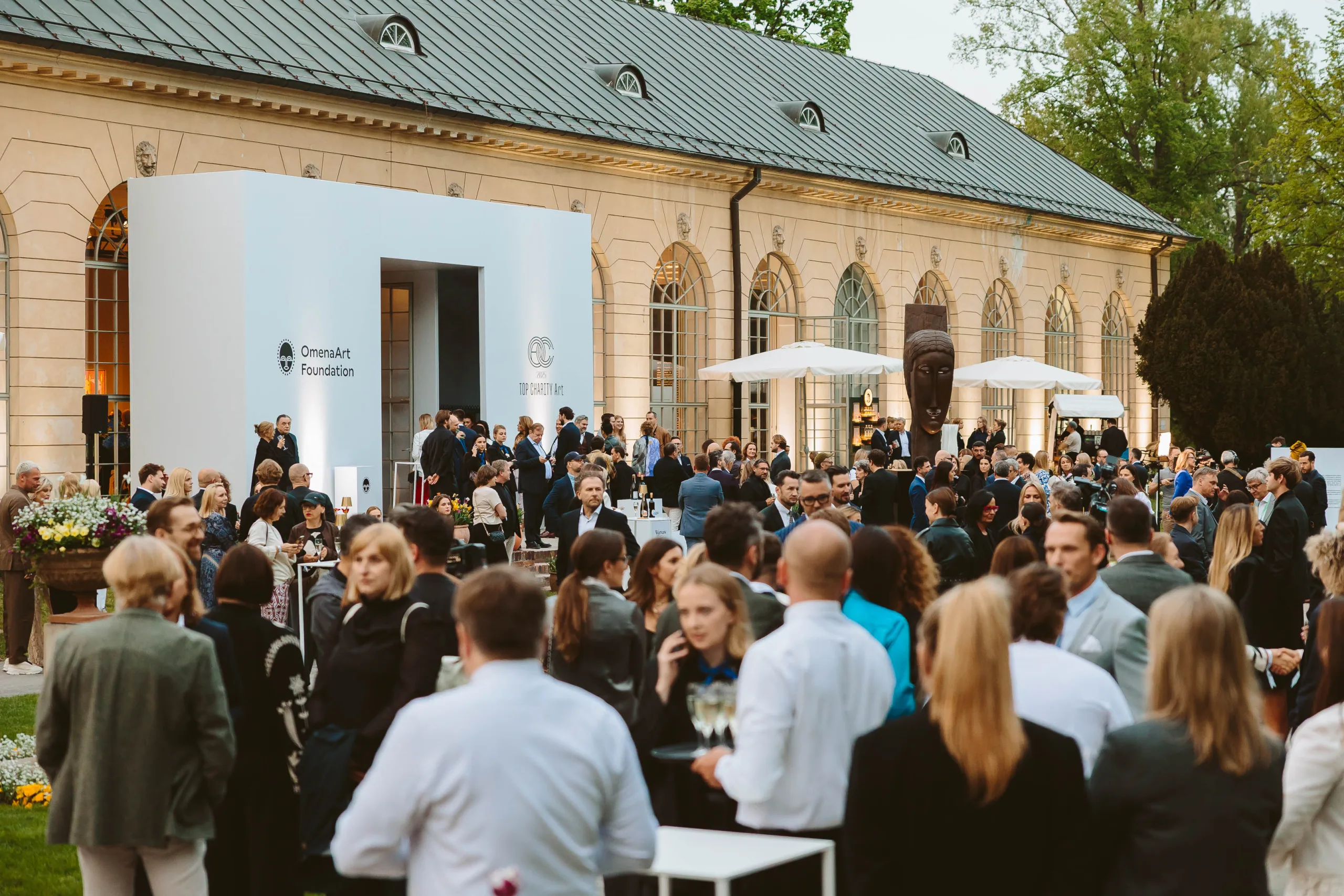 Guests at a garden reception in front of a historic building with arched windows, a white entrance and parasols in the background