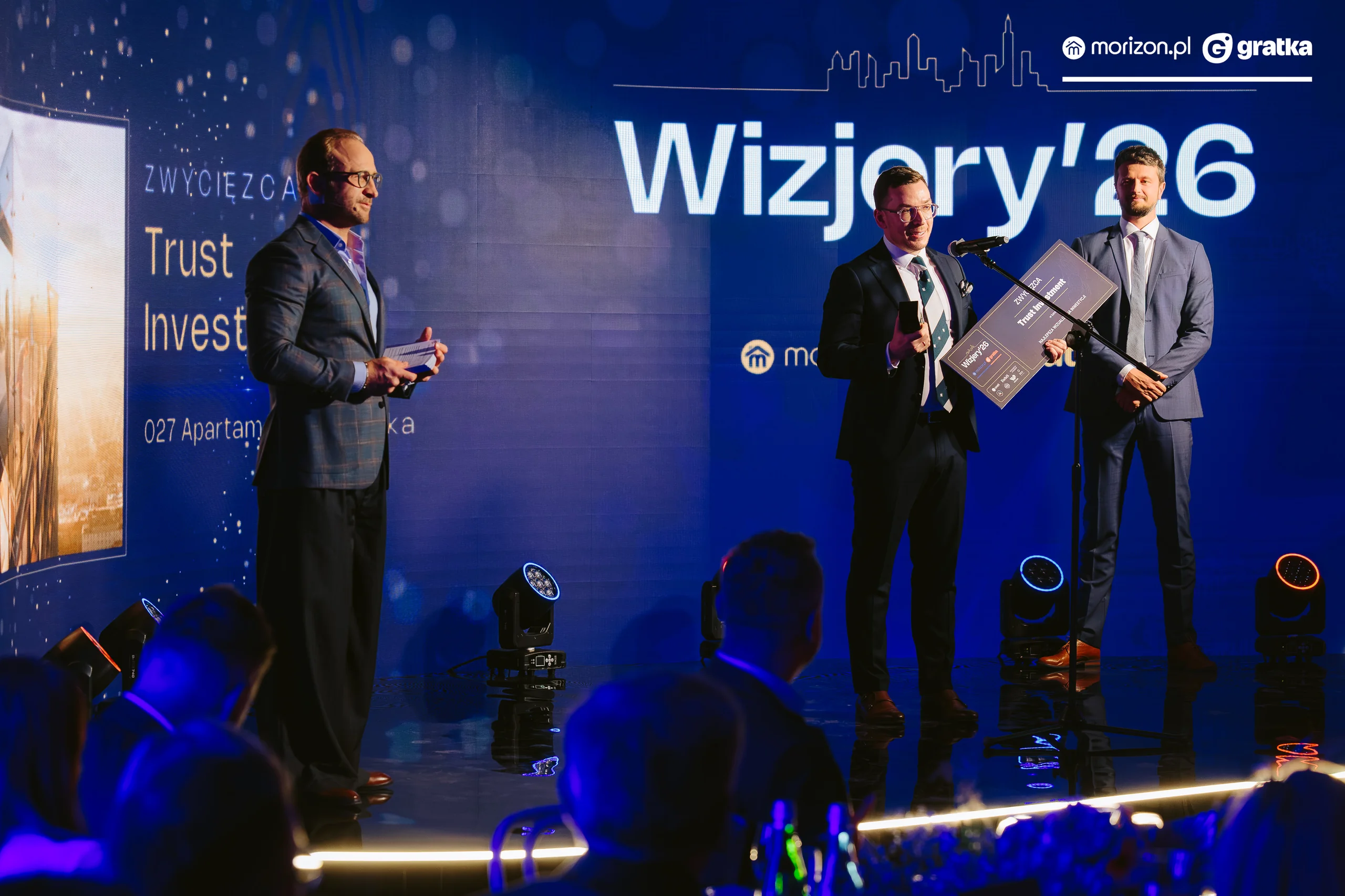 The category winner, holding their certificate, gives a speech at the microphone on the stage at the awards gala, with the host and guests at the tables in the foreground